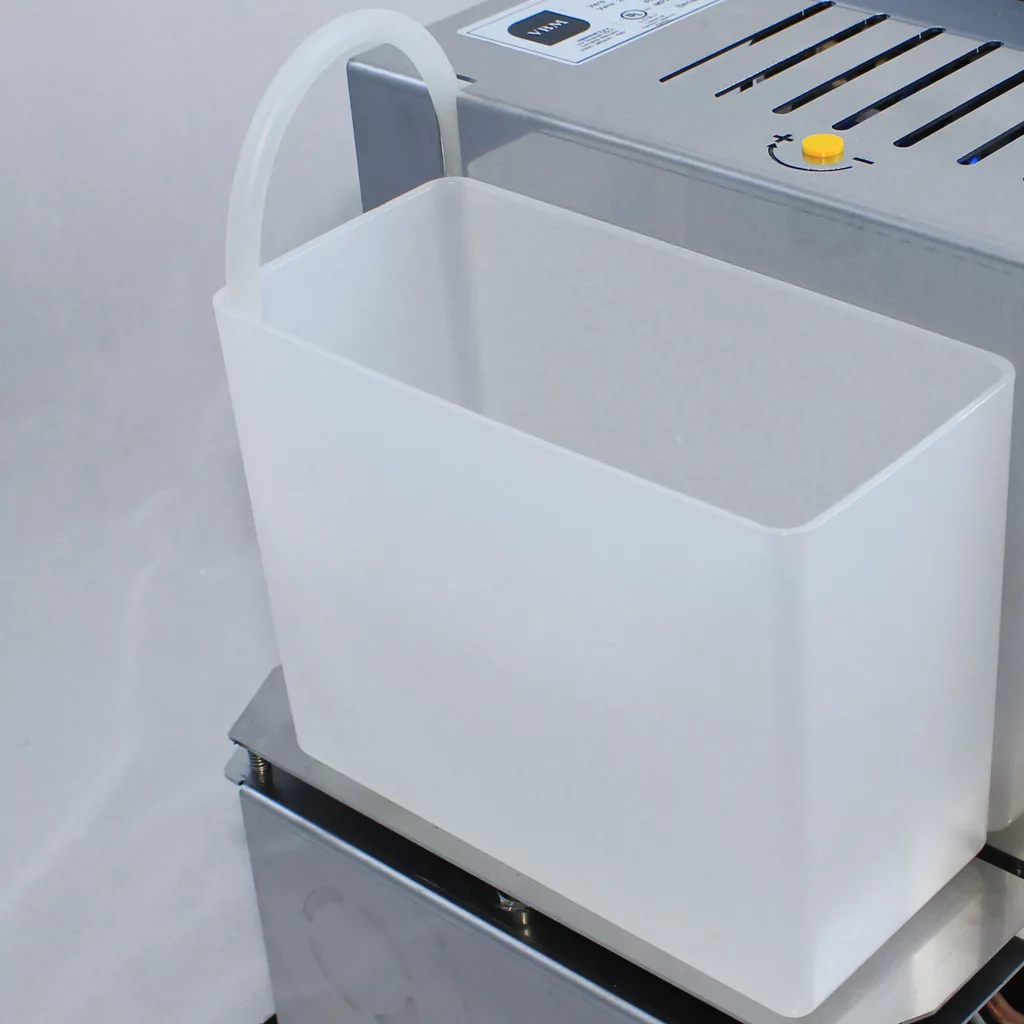 A translucent rectangular plastic container with a curved tube sits next to a DISCONTINUED VBM Domobar Super V4 Dual Boiler PID Switchable Espresso Machine, which features buttons and vents. © 2025 1st-line.com