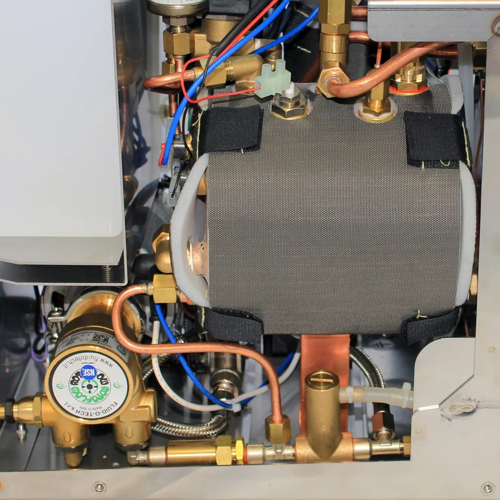 Close-up of the internal components of the discontinued VBM Domobar Super V4 Dual Boiler PID Switchable Espresso Machine, showing pipes, wires, a pressure gauge, and an insulated cylinder within a compact assembly. © 2025 1st-line.com