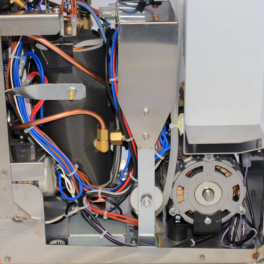 Close-up view of the internal components of the discontinued VBM Domobar Super V4 Dual Boiler PID Switchable Espresso Machine, featuring copper pipes, colored wires, a motor, and a black cylinder within its metal frame. © 2025 1st-line.com