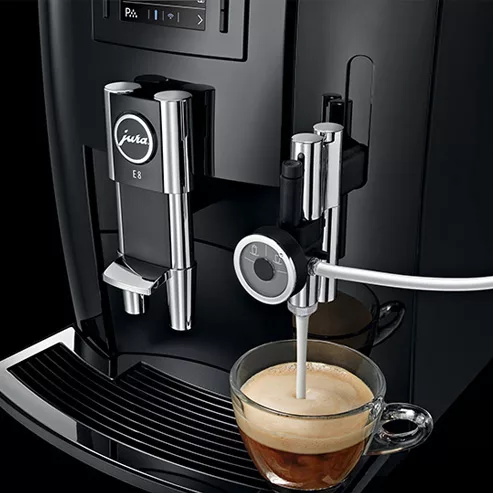 The DISCONTINUED Jura E8 Super-Automatic Coffee Machine uses a milk hose to automatically froth and dispense milk into a glass cup of coffee on its drip tray. © 2025 1st-line.com