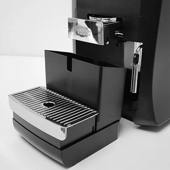 A close-up black and white image shows the REDIRECTED Ascaso Kap Tronic Capsule Espresso Machine in black, with its drip tray and grounds container pulled out to reveal the metal grid and plastic compartments. © 2025 1st-line.com
