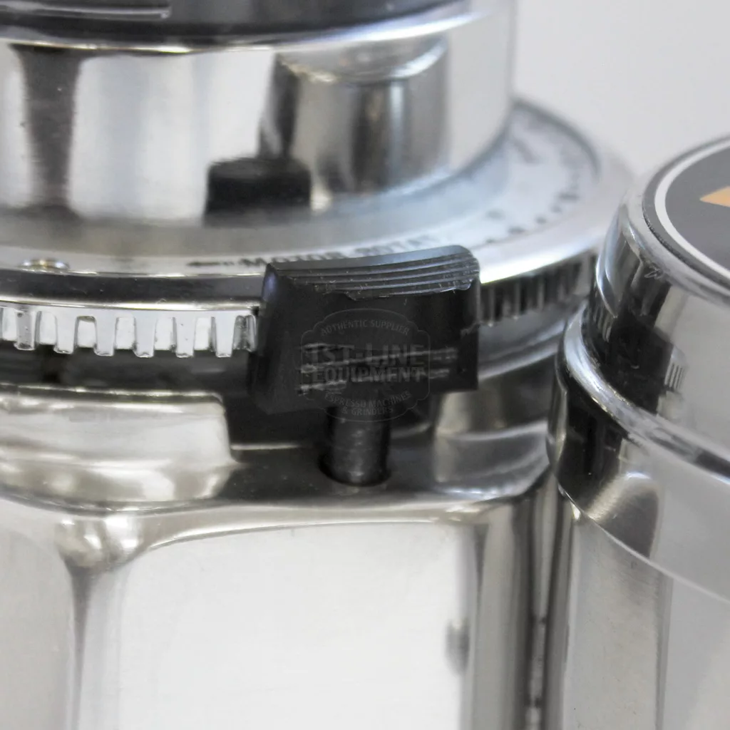 Close-up of the REDIRECTED Quamar Q50 Commercial Doserless Espresso Coffee Grinder - electronic, chrome, showing its shiny stainless steel body and a black plastic textured latch with engraved text on the side. © 2025 1st-line.com