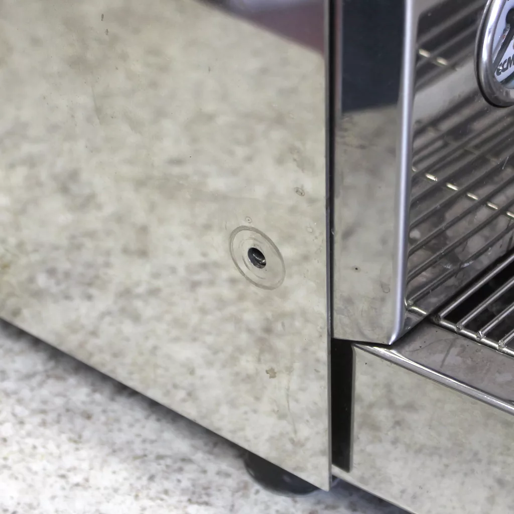 A close-up of a (SOLD) SIDEWALK SALE - ECM Veronica Espresso Machine USED E002 E502 shows a small dent in its reflective stainless steel body, with a vented section and visible screw, sitting on a speckled countertop. © 2025 1st-line.com