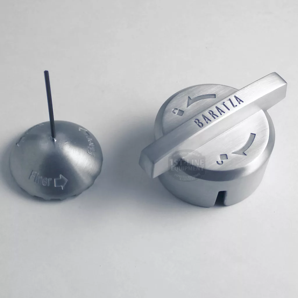 Two metallic parts from the DISCONTINUED Baratza Vario Coffee Grinder are shown on a white background: one is a round piece with a thin rod labeled “Finer,” and the other is a round cap with a handle engraved “BARATZA.”. © 2025 1st-line.com