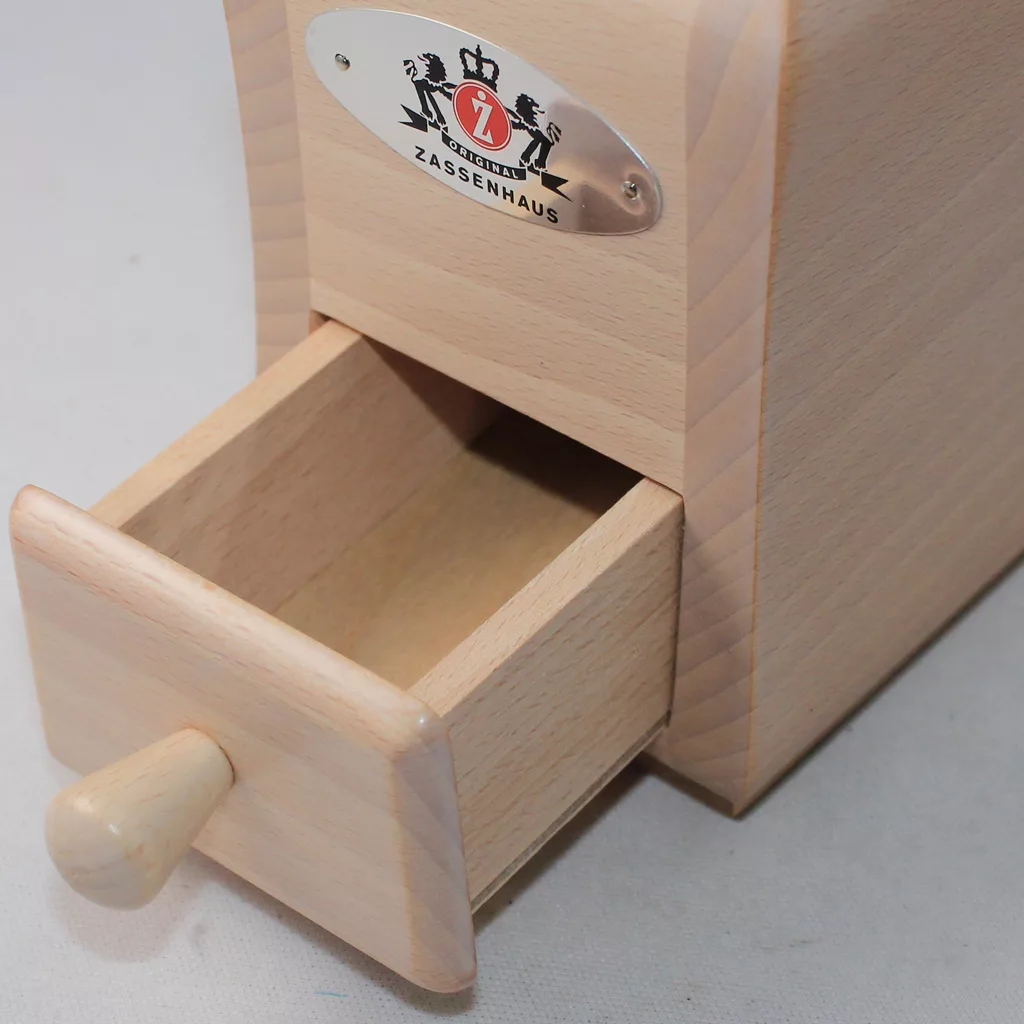 A close-up of the DISCONTINUED Zassenhaus Santiago Hand Coffee Grinder / Coffee Mill in varnished beech, showing its open small drawer with light wood inside and a metal brand label on the front. © 2025 1st-line.com