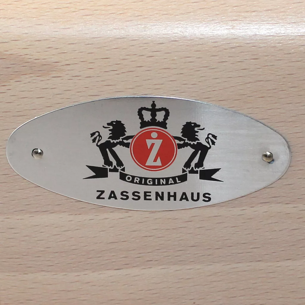 A metallic oval Zassenhaus logo plate with two lions, a crown, and a red Z emblem is mounted with two screws on the varnished beech surface of the discontinued Zassenhaus Santiago Hand Coffee Grinder / Coffee Mill. © 2025 1st-line.com