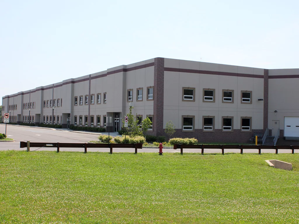 A large, modern industrial warehouse building with multiple windows and loading docks, set beside a road with signs and surrounded by green grass and a fire hydrant in the foreground. © 2025 1st-line.com