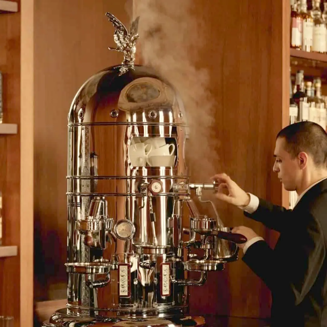 A barista in a suit uses the Elektra Belle Epoque 2 Group Commercial Espresso Machine, featuring an eagle on top and ornate design, as it releases steam inside a wooden-paneled, bottle-lined room.
