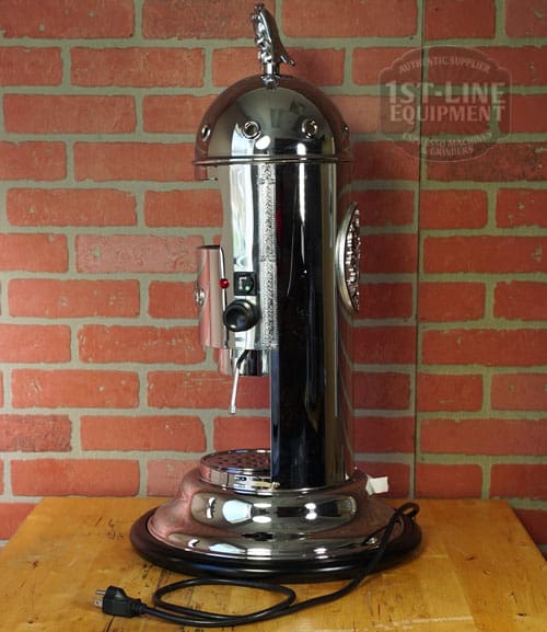 A chrome SIDEWALK SALE - Elektra Mini Verticale A1C Espresso Machine (E211) with a cylindrical body and domed top sits on a wooden surface against a red brick wall, its coiled power cord at the base beside the lever and control button. © 2025 1st-line.com