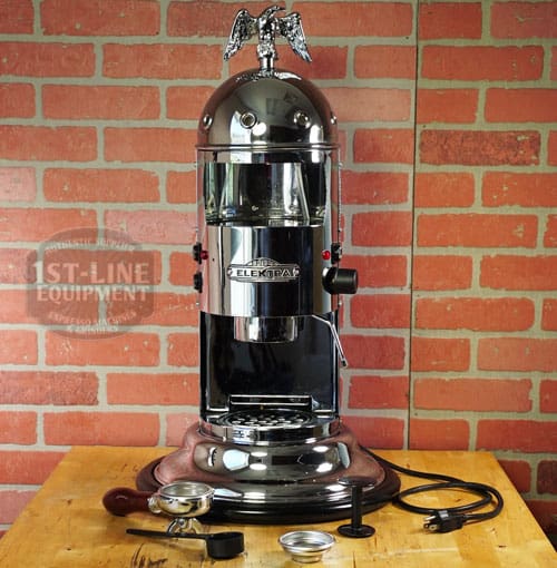 A chrome SIDEWALK SALE - Elektra Mini Verticale A1C Espresso Machine (E211) with a vintage eagle topper sits on a wooden table by a brick wall, paired with coffee accessories and power cord—your perfect Sidewalk Sale addition. © 2025 1st-line.com