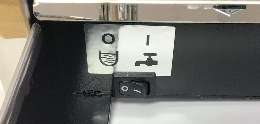 A close-up of a black switch on the DISCONTINUED ECM Elektronika II Profi HX Espresso Machine shows two symbols below it: a cup with wavy lines labeled O and a faucet labeled I, indicating different water functions. © 2025 1st-line.com