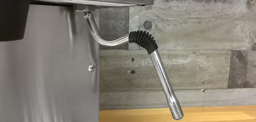 A close-up of a metal steam wand with a black rubber grip on the Rancilio Silvia M Espresso Machine, set against a wooden and metal background. © 2025 1st-line.com