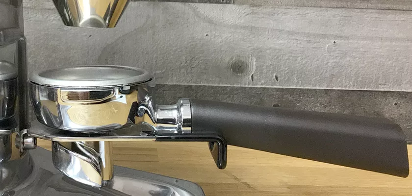 A close-up of a portafilter with a black handle attached to a coffee machine on a wooden countertop, with a DISCONTINUED Quamar M80E V2 On Demand Espresso Coffee Grinder in the background against a gray wooden wall. © 2025 1st-line.com