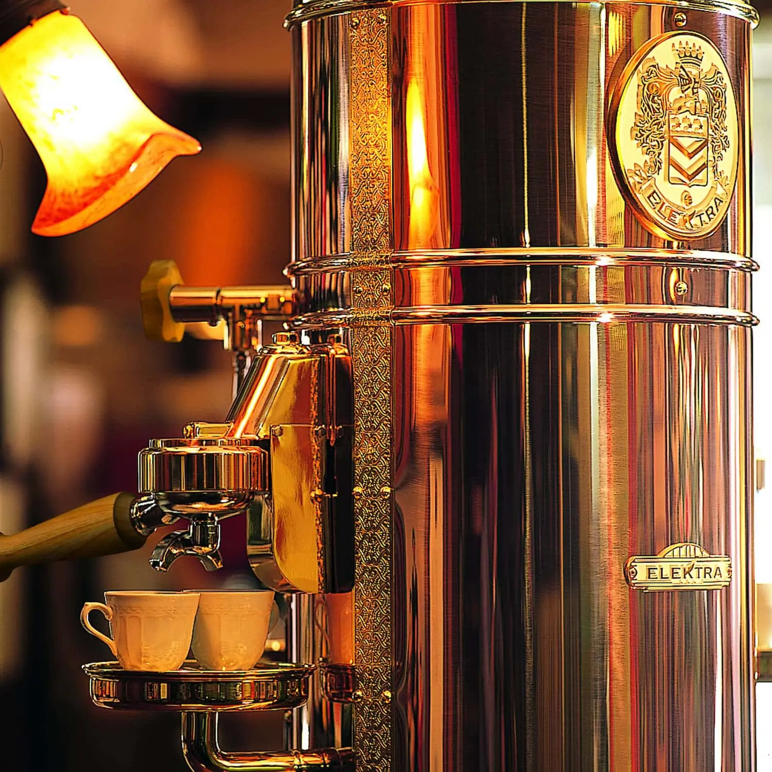 A close-up of the Elektra Belle Epoque 2 Group Commercial Espresso Machine, featuring shiny vintage details, as it prepares two cups of espresso beneath a warm orange lamp.
