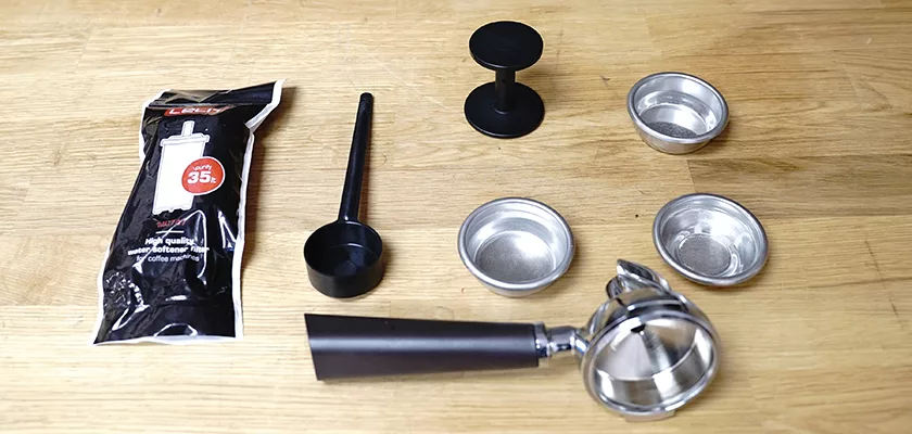 A collection of espresso accessories on wood, including a tamper, scoop, two metal filter baskets, portafilter, and ground coffee for the DISCONTINUED Lelit Victoria PL91T PID Single Boiler Espresso Machine. © 2025 1st-line.com