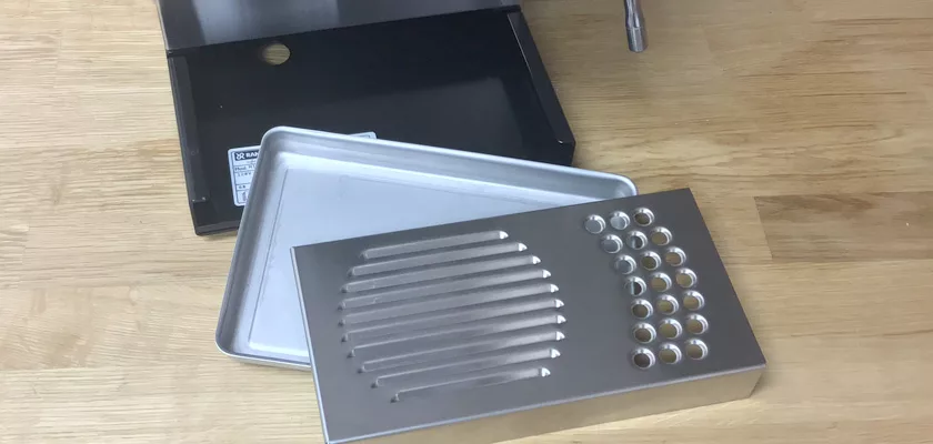 A disassembled metal drip tray for the Rancilio Silvia M Espresso Machine is displayed on a wooden surface, showing the outer box, inner tray, and slotted cover with drainage holes. © 2025 1st-line.com