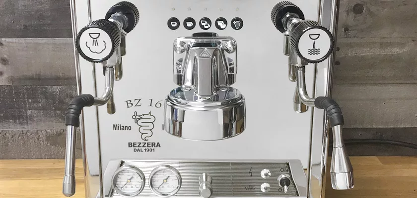 A discontinued Bezzera BZ16 DE HX Espresso Machine with two steam wands, pressure gauges, and control knobs sits on a wooden countertop against a rustic wood wall. © 2025 1st-line.com