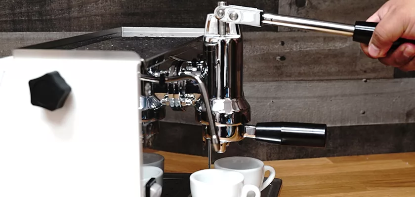 A hand operates the lever of a DISCONTINUED Ponte Vecchio Lusso 1 Group Spring Piston Lever Espresso Machine, brewing espresso into two small white cups on a wooden counter with a rustic wood wall in the background. © 2025 1st-line.com