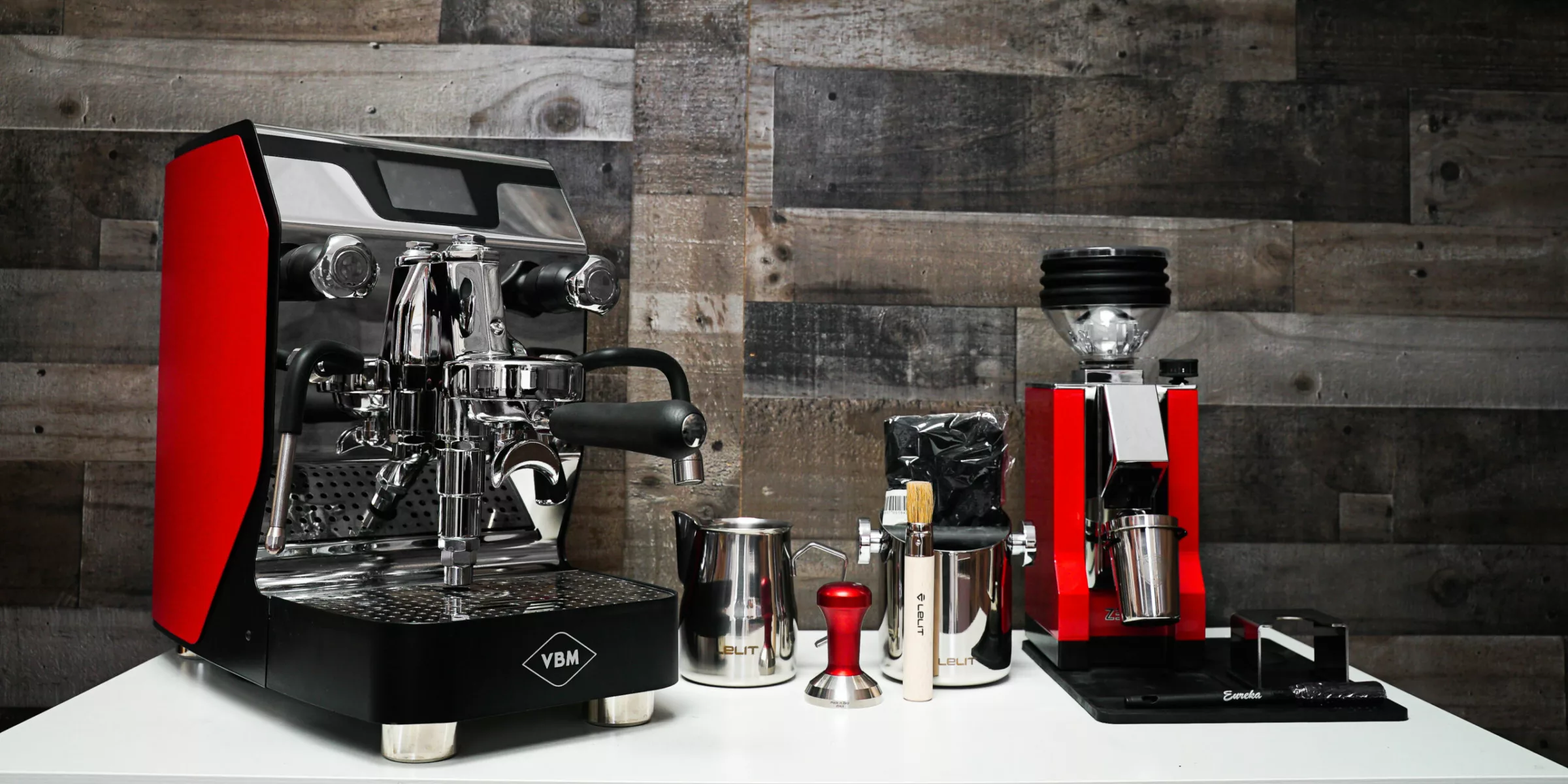 A home coffee bar on a white counter features the Build My VBM Espresso Bar, a red coffee grinder, metal milk pitchers, a tamper, brush, and black bag of beans—all set against a rustic wood wall. © 2025 1st-line.com