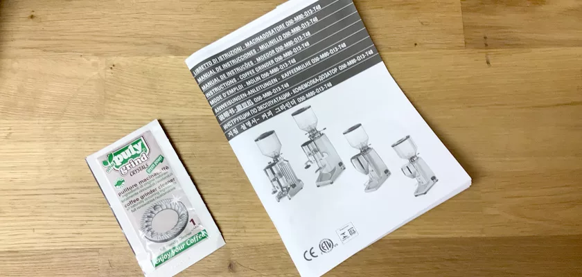 A packet of SantAnna instant yeast rests on a wooden surface beside a manual that displays images and multilingual details for the DISCONTINUED Quamar M80E V2 On Demand Espresso Coffee Grinder. © 2025 1st-line.com