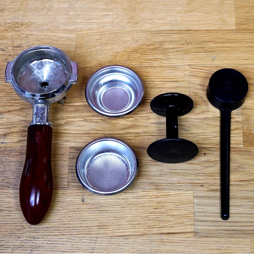 A portafilter with a wooden handle, two metal filter baskets, a black coffee tamper, and dosing tool—essential accessories to pair with the SIDEWALK SALE - Elektra Mini Verticale A1C Espresso Machine - Chrome (E211). © 2025 1st-line.com