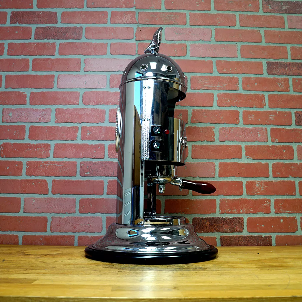A SIDEWALK SALE - Elektra Mini Verticale A1C Espresso Machine - Chrome (E211) shines on a wooden countertop, set against a red brick wall. © 2025 1st-line.com
