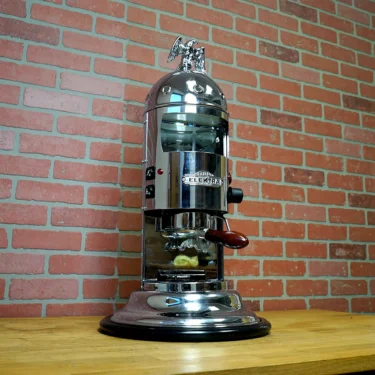 A SIDEWALK SALE - Elektra Mini Verticale A1C Espresso Machine - Chrome (E211) with a dome top and eagle ornament is displayed on a wooden counter against a red brick wall. © 2025 1st-line.com