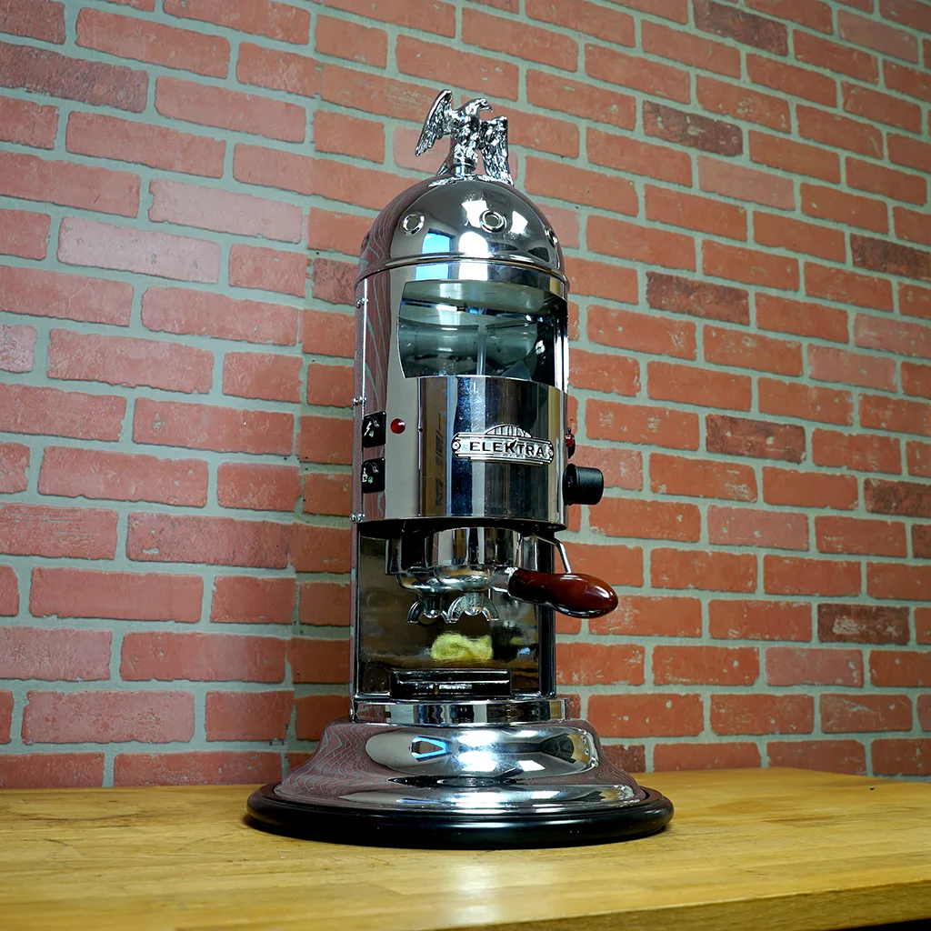 A SIDEWALK SALE - Elektra Mini Verticale A1C Espresso Machine - Chrome (E211) with a dome top and eagle ornament is displayed on a wooden counter against a red brick wall. © 2025 1st-line.com