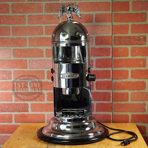 A SIDEWALK SALE - Elektra Mini Verticale A1C Espresso Machine - Chrome (E211) with an eagle finial stands on a wooden table by a red brick wall, the power cord neatly coiled beside its base. © 2025 1st-line.com