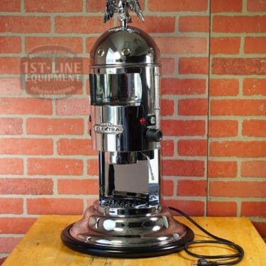 A SIDEWALK SALE - Elektra Mini Verticale A1C Espresso Machine - Chrome (E211) with an eagle ornament sits on a round base before a red brick wall, its unplugged power cord coiled neatly to the side. © 2025 1st-line.com