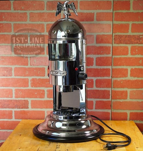 A SIDEWALK SALE - Elektra Mini Verticale A1C Espresso Machine - Chrome (E211) with an eagle ornament sits on a round base before a red brick wall, its unplugged power cord coiled neatly to the side. © 2025 1st-line.com