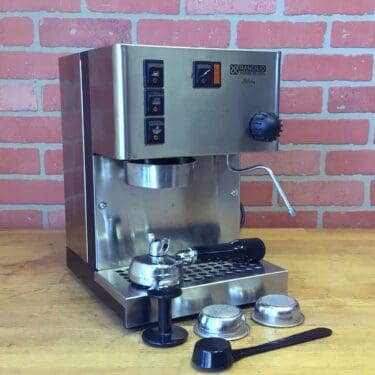 A SIDEWALK SALE - Rancilio Silvia Espresso Machine (USED) (E113) sits on a wooden counter before a pink brick wall, with its portafilter, tamper, filter baskets, and coffee scoop neatly arranged in front. © 2025 1st-line.com