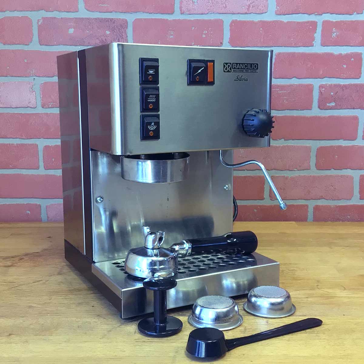 A SIDEWALK SALE - Rancilio Silvia Espresso Machine (USED) (E113) sits on a wooden counter before a pink brick wall, with its portafilter, tamper, filter baskets, and coffee scoop neatly arranged in front. © 2025 1st-line.com