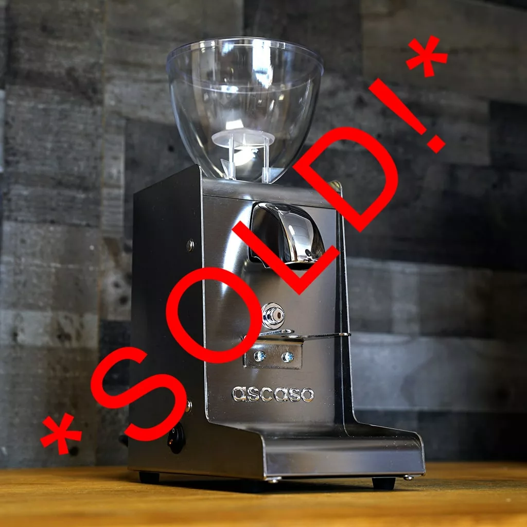A stainless steel Ascaso I-Steel Conical Burr Espresso Grinder (2018 OPEN BOX DISPLAY) sits on a wooden surface with a clear hopper, against a gray wood panel wall. Large red SOLD! text marks it as part of our SIDEWALK SALE. © 2025 1st-line.com