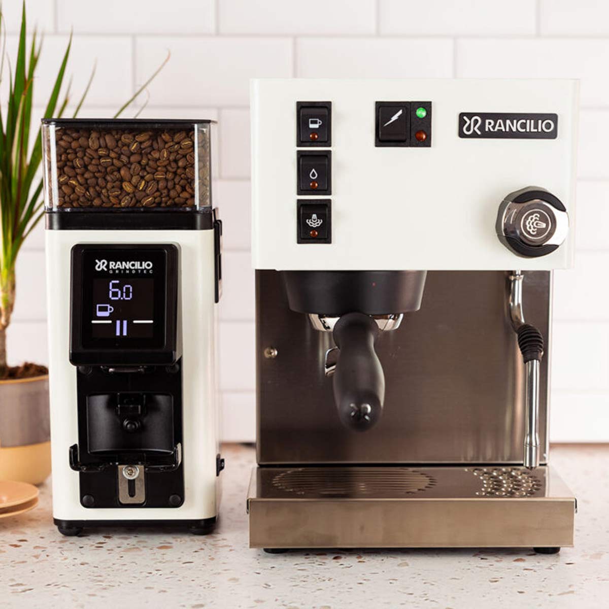 A white Rancilio Silvia M Espresso Machine and a matching grinder with digital display sit on the kitchen counter, coffee beans prepped in the grinder and a small plant in the background. © 2025 1st-line.com
