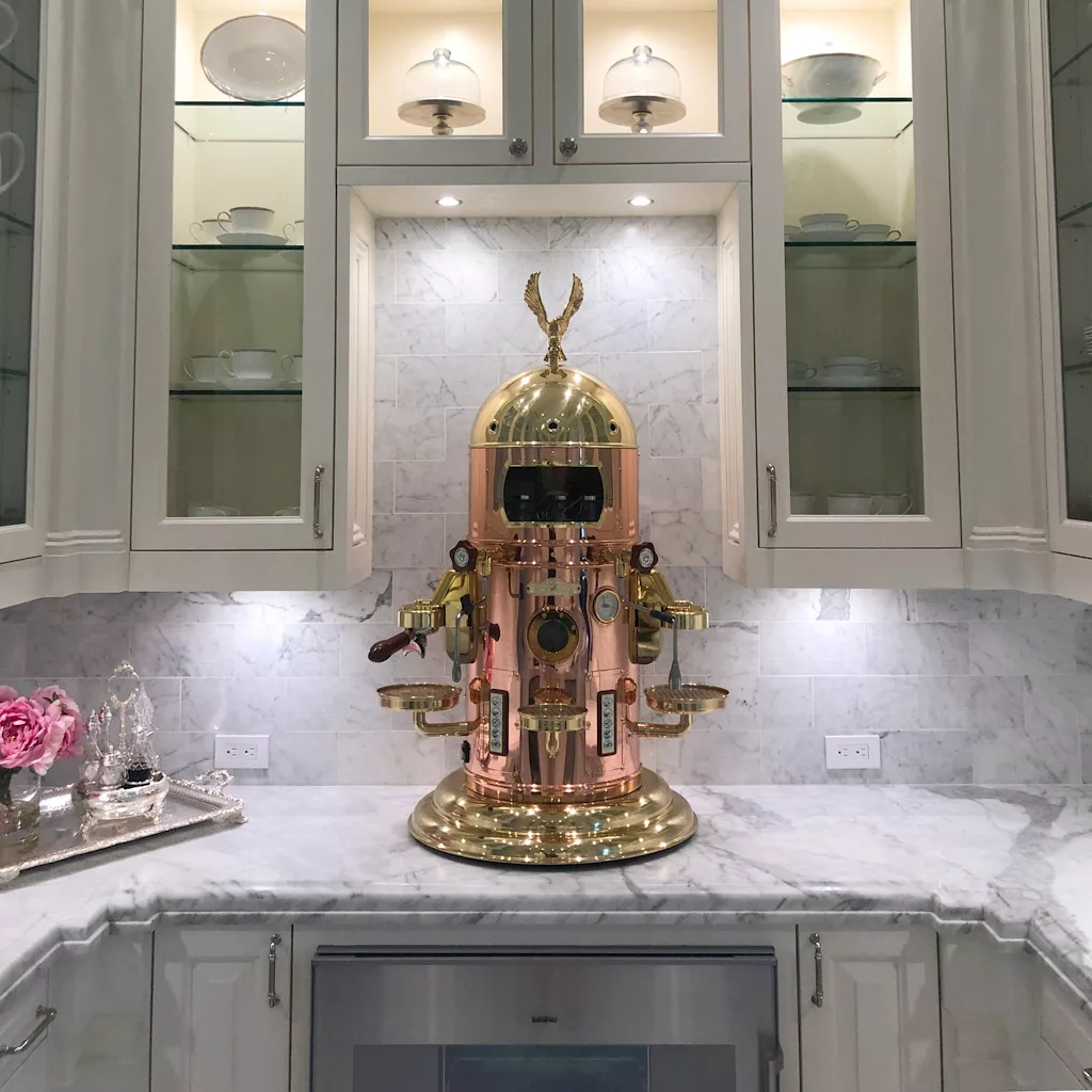 An Elektra Belle Epoque 2 Group Commercial Espresso Machine sits on a marble counter, framed by glass-front cabinets filled with white dishes and illuminated by warm overhead lighting. © 2025 1st-line.com