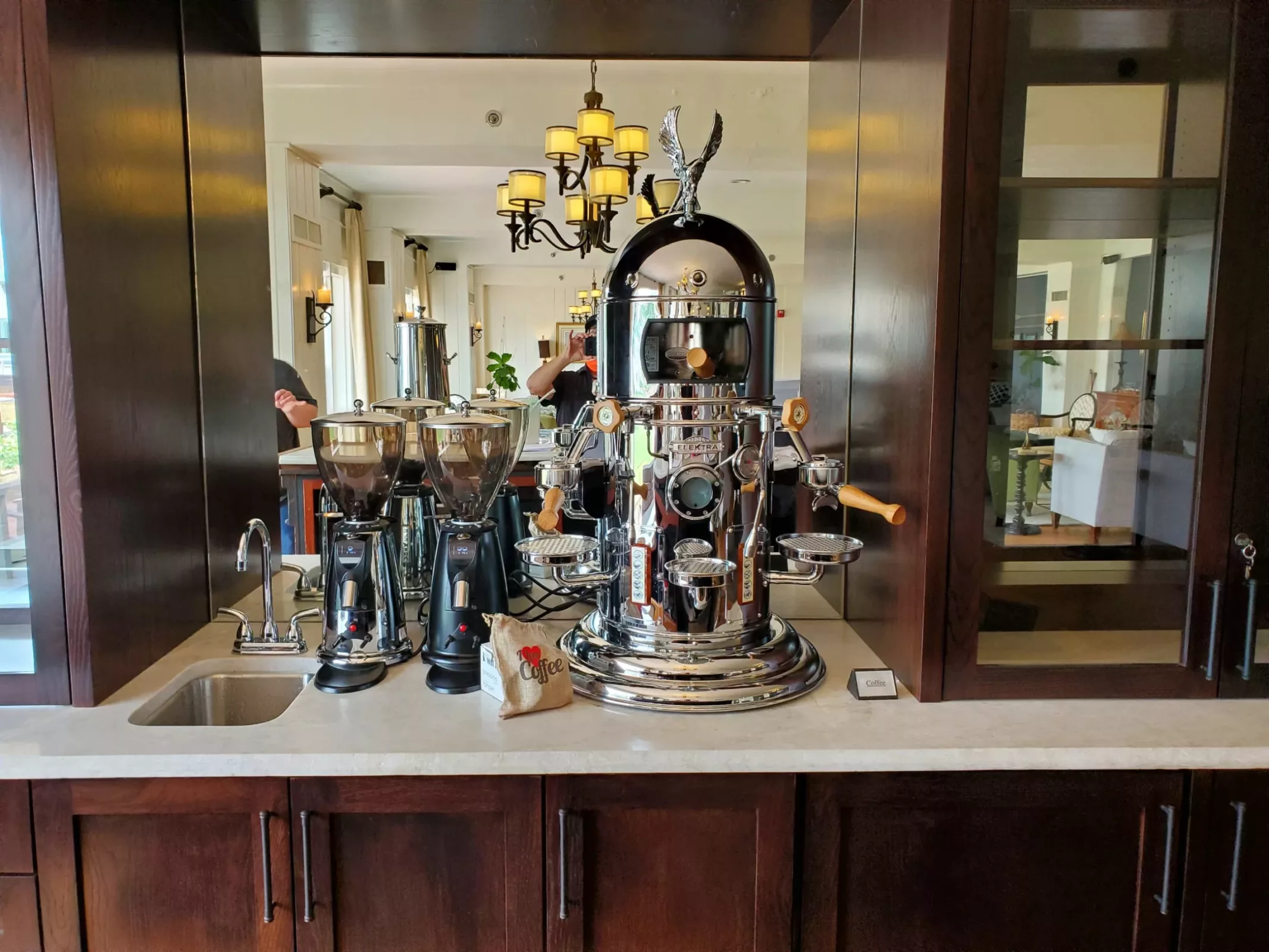 An Elektra Belle Epoque 2 Group Commercial Espresso Machine with wooden handles sits on a countertop next to two coffee grinders and a bag of coffee, surrounded by dark wood cabinets and warm lighting in a cozy café. © 2025 1st-line.com