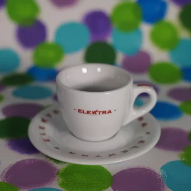An Elektra TE Espresso Cup & Saucer in white rests on a surface, set against a backdrop of blue, green, and purple polka dots. © 2025 1st-line.com