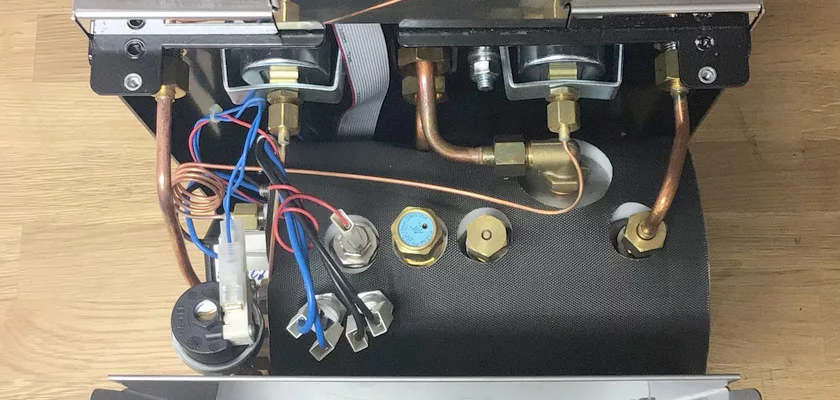An open DISCONTINUED ECM Elektronika II Profi HX Espresso Machine displays its internal copper pipes, wiring, connectors, and metal fittings on a black base set against a wooden surface. © 2025 1st-line.com