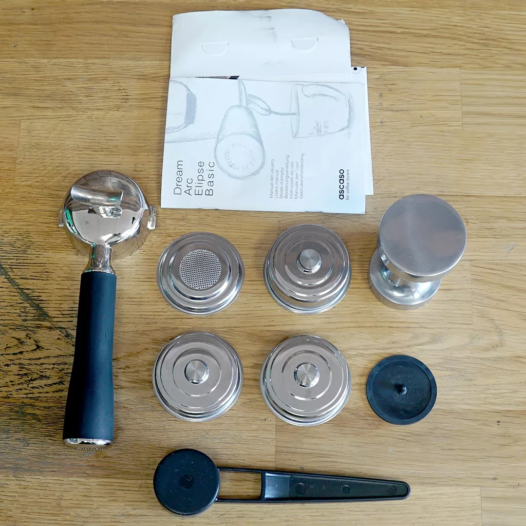 Assorted parts for the Ascaso Dream V1 UP Meadow Green Espresso Machine Tank/Reservoir (USED)—including portafilter, filter baskets, tamper, tools, and manual—are displayed on a wooden surface. Ideal for upgrading your used setup. © 2025 1st-line.com