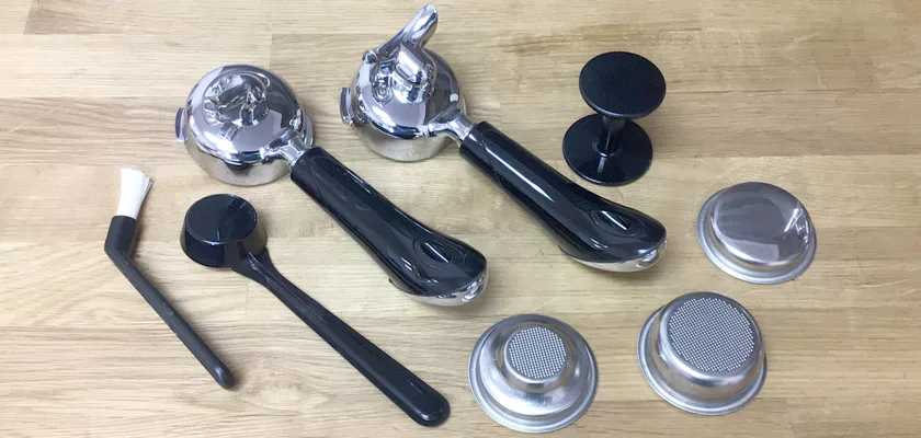 Barista tools on a wooden surface—two portafilters, two black tampers, a coffee grounds brush, and metal filter baskets—perfect accessories for your DISCONTINUED Bezzera BZ16 DE HX Espresso Machine. © 2025 1st-line.com