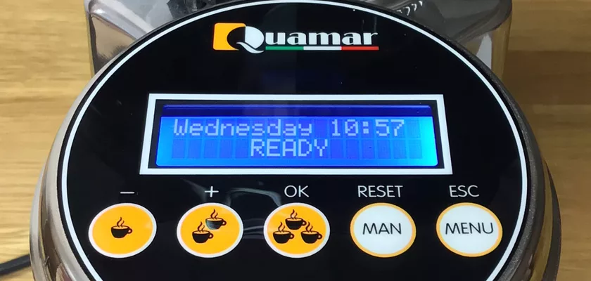 Close-up of a DISCONTINUED Quamar M80E V2 On Demand Espresso Coffee Grinder display showing Wednesday 10:57 READY, with menu, manual, reset, OK, and coffee cup buttons. The background is a wooden surface. © 2025 1st-line.com