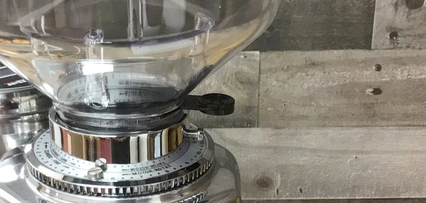 Close-up of a DISCONTINUED Quamar M80E V2 On Demand Espresso Coffee Grinder with a clear hopper and metal adjustment ring, partially visible at the left against a wooden plank background. © 2025 1st-line.com