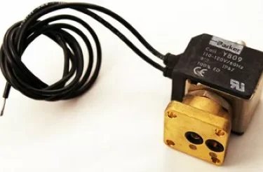 Close-up of a VBM UNK61559532 3-Way Solenoid Valve with a black coil, electrical wires, and a rectangular brass base featuring two circular openings. © 2025 1st-line.com