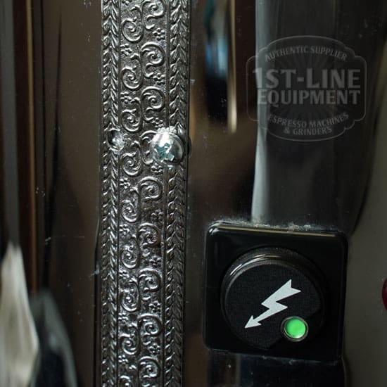 Close-up of the chrome Elektra Mini Verticale A1C Espresso Machine (E211), showing a black button with a white lightning bolt and green indicator light. The 1st-Line Equipment logo is reflected in its polished finish. © 2025 1st-line.com