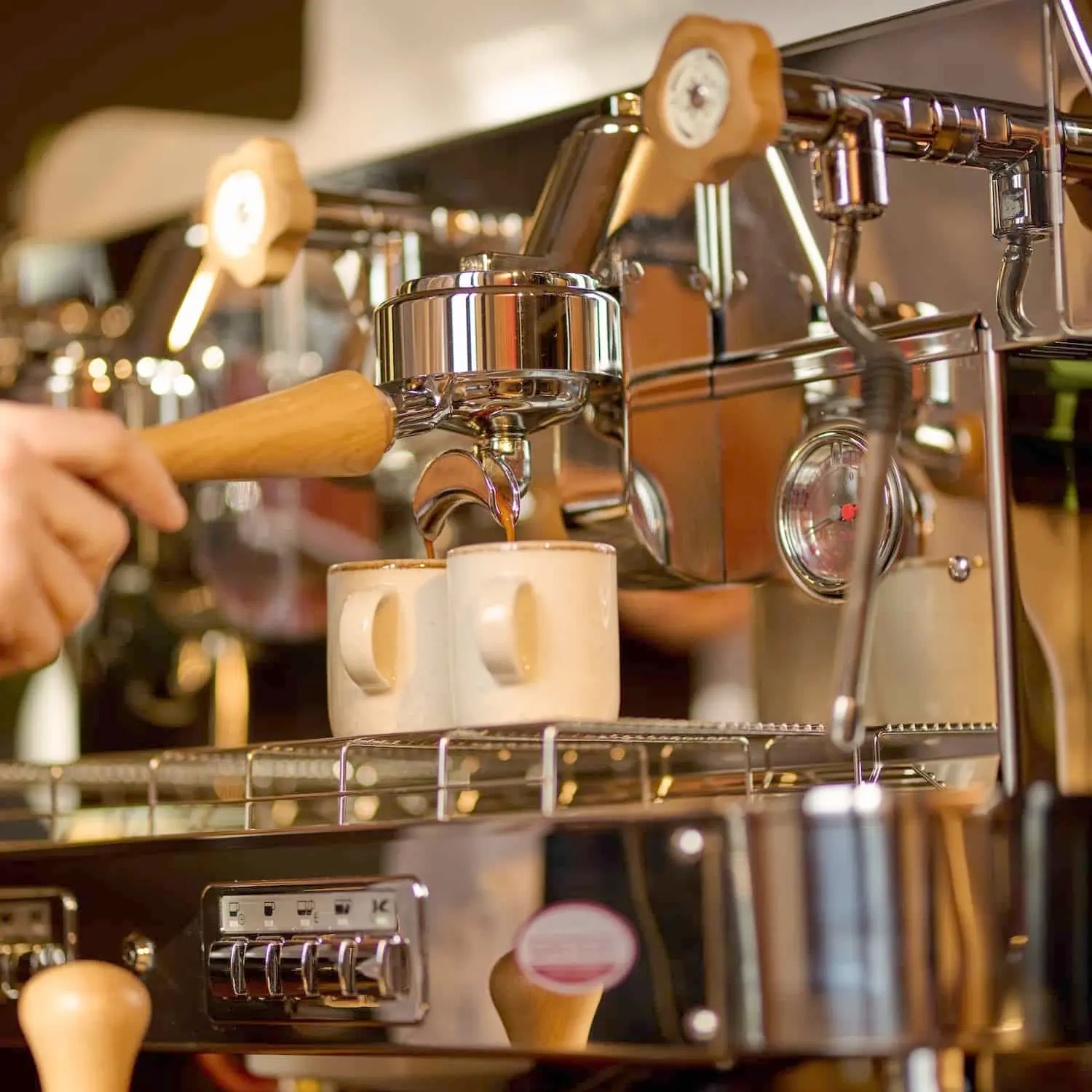Close-up of the Elektra Barlume 2 Group Commercial Espresso Machine brewing coffee into two white mugs, with a hand gripping the portafilter, creating a warm, inviting café ambiance.