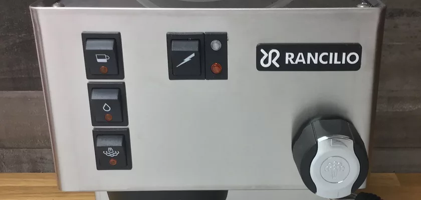 Close-up of the Rancilio Silvia M Espresso Machine control panel with three square buttons for beverage, water, and steam, a lightning bolt switch, indicator lights, and a large black knob on the right. © 2025 1st-line.com