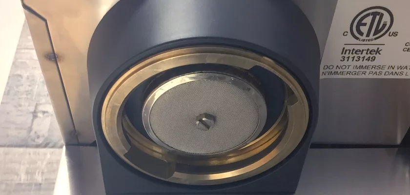 Close-up of the Rancilio Silvia M Espresso Machine group head with a circular metal filter and brass ring, next to a silver label displaying certification marks and instructions. © 2025 1st-line.com