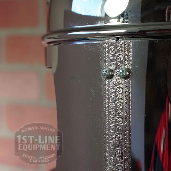 Close-up of the SIDEWALK SALE - Elektra Mini Verticale A1C Espresso Machine - Chrome (E211) with ornate engraving and screws; a blurred brick wall and translucent 1st-Line Equipment logo appear in the background. © 2025 1st-line.com
