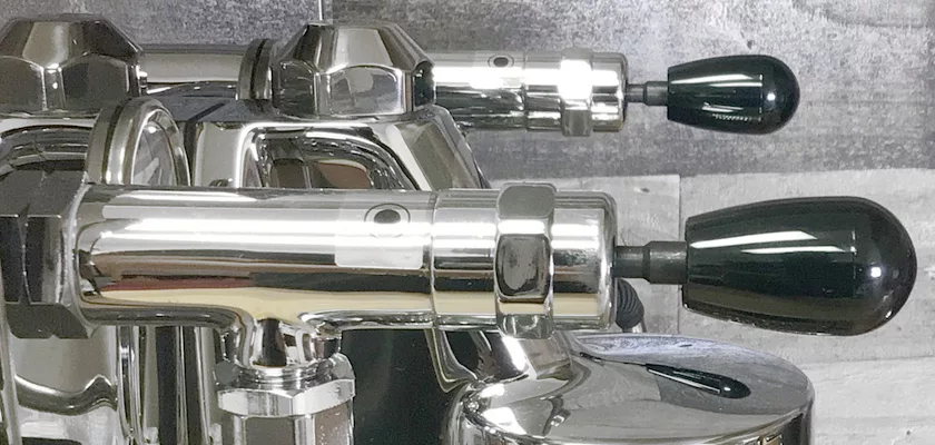 Close-up of two chrome levers with black handles on the DISCONTINUED ECM Elektronika II Profi HX Espresso Machine, set against a gray and silver textured background. The polished metal surfaces reflect light and nearby details. © 2025 1st-line.com